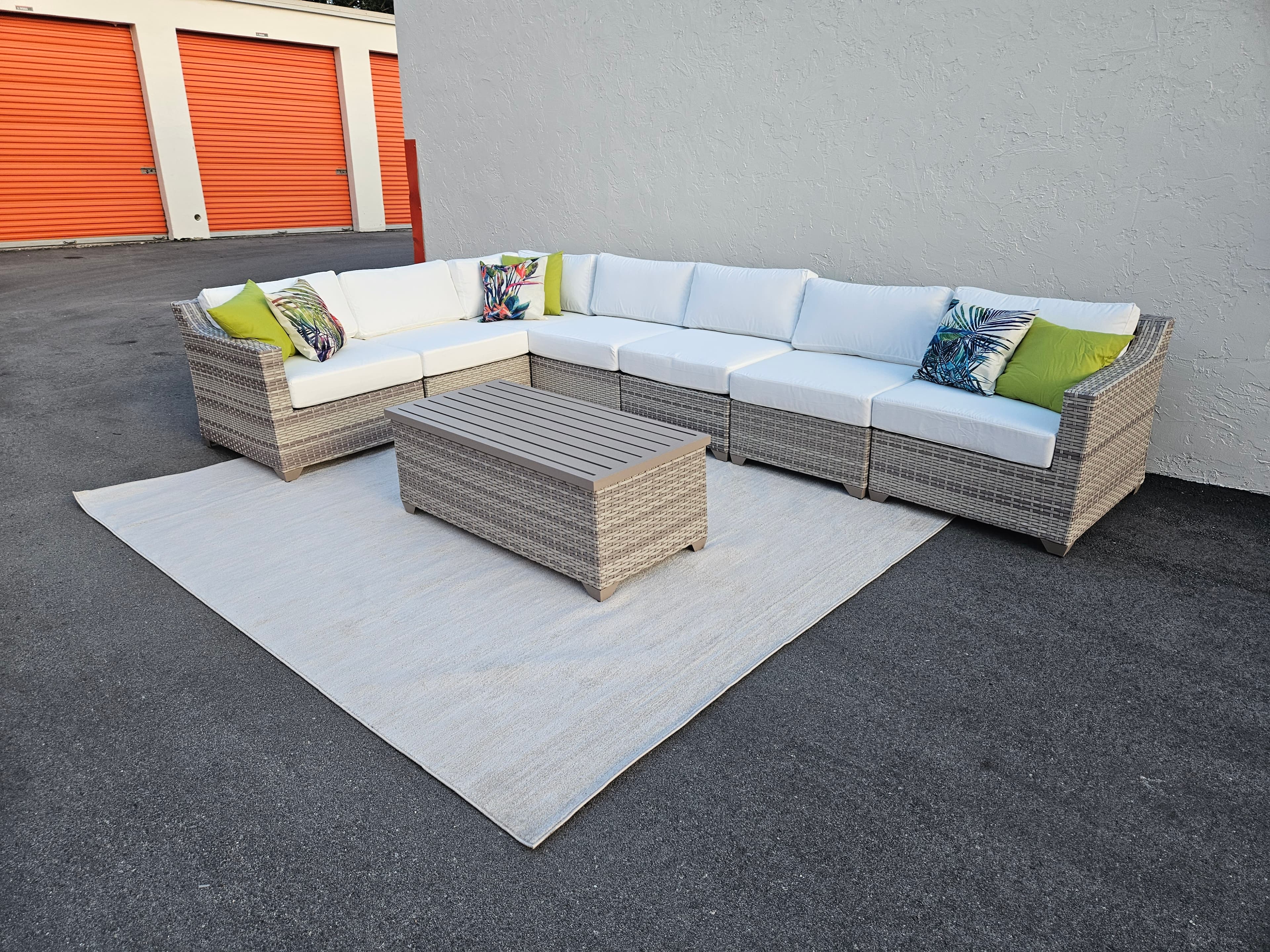 Grey wicker outdoor sectional with white cushions and tropical pillows on a grey rug.